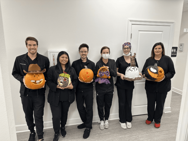Dental team members standing in a line holding their carved pumpkins while smiling at the camera.