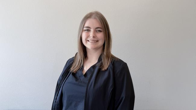 A person smiling while standing up against a blank wall